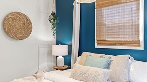 A bedroom with a bed, a bedside table with a lamp, a hanging plant, and a window with blinds.