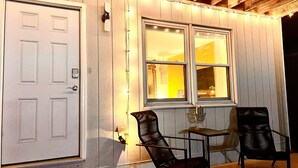 A white door with a keyhole and a small mailbox, a window with a view of a lamp and a plant inside, and two black chairs with a small table in front.
