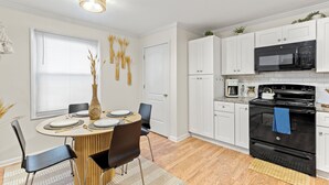 A modern kitchen with white cabinets, a black stove, and a microwave. There is a dining table with chairs and a vase with dried plants.