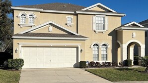 A two-story house with a beige exterior, a white garage door, and a small front yard with a sidewalk leading to the entrance.