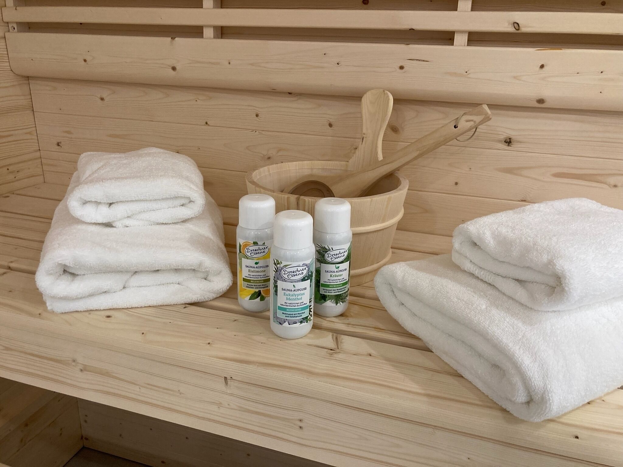 Un banc en bois sur lequel sont pliées des serviettes blanches, deux bouteilles de Schwarze Essenz et un seau en bois avec une cuillère en bois.