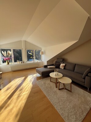 En moderne stue i loftstil med højt til loftet, store vinduer, en grå sofa, to runde sofaborde og en hvid måtte.
