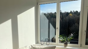 Una sala de estar moderna con un sofá color beige, un ventanal con vista a árboles y una planta en maceta en el alféizar.