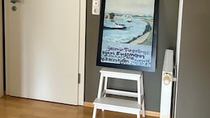 Una habitación con una puerta blanca, un cuadro enmarcado de una escena costera, un taburete blanco pequeño y un radiador blanco.