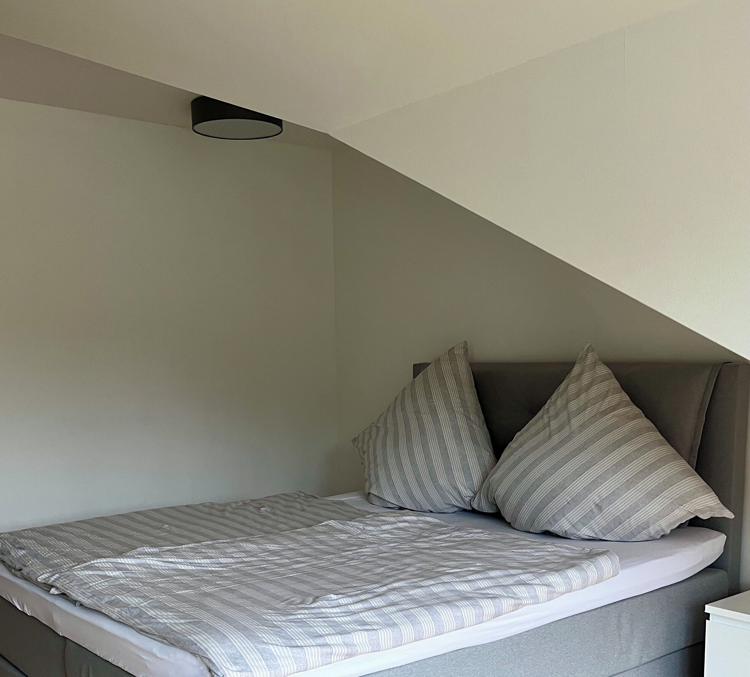 Une petite chambre à coucher moderne avec un plafond en pente, un lit simple avec une literie à rayures et deux oreillers.