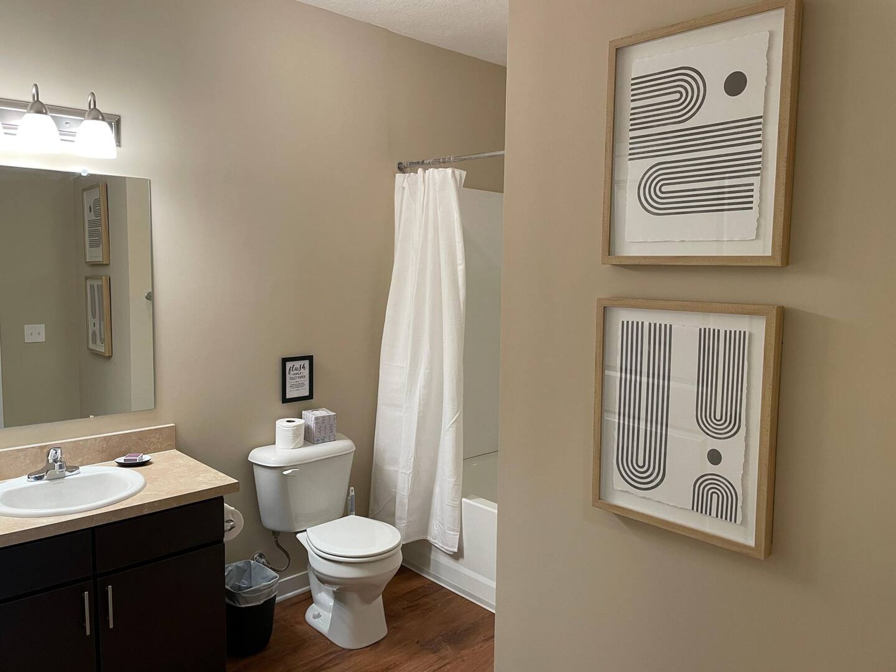 Ein Badezimmer mit Waschbecken, Toilette und Duschvorhang. An der Wand hängen gerahmte Kunstwerke.