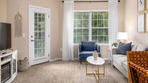 A living room with a TV, a blue chair, a white table, a sofa, and a window with blinds.
