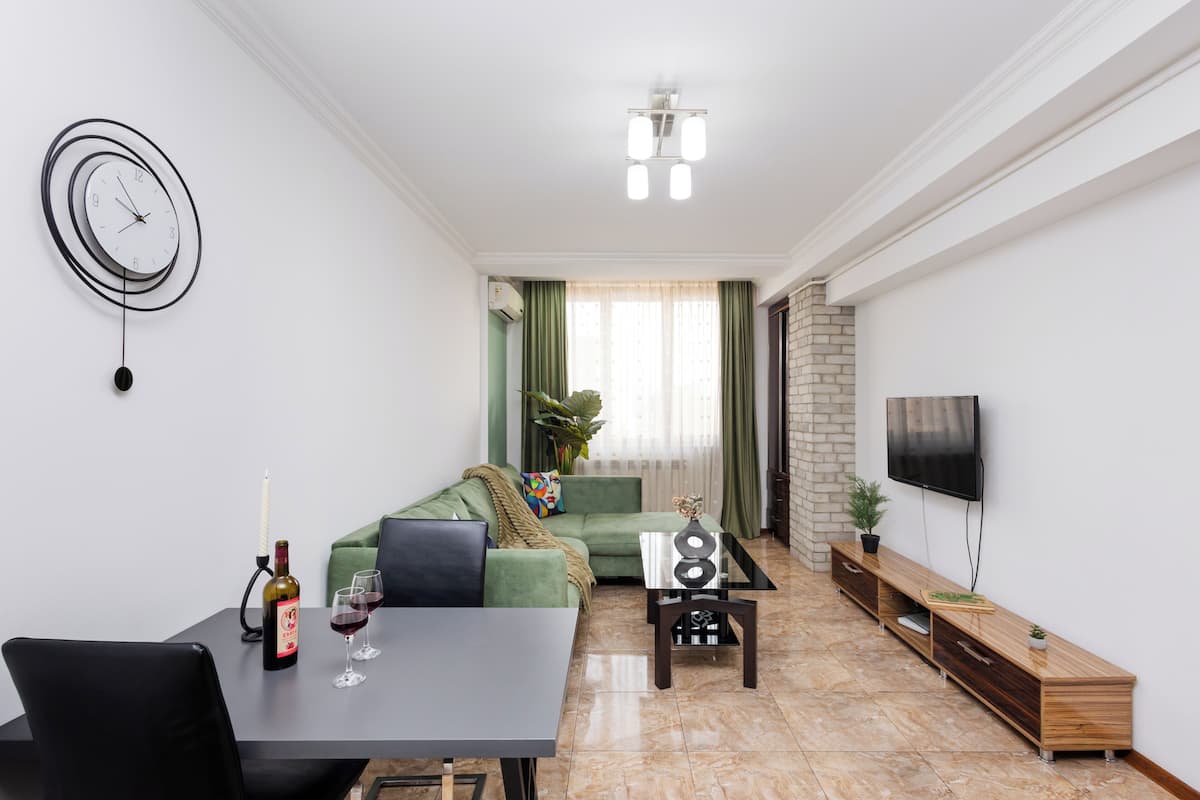 A modern living room with a sofa, a dining table with wine, a TV, and a unique wall clock.