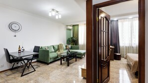 A modern living room with a green sofa, a glass coffee table, and a dining table with chairs. There is a bed with a wooden headboard in the background.