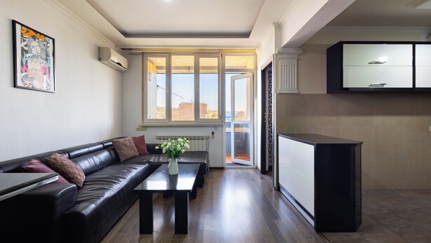 A modern living room with a black leather sofa, a coffee table with a vase of flowers, and a view through large windows to the outside.