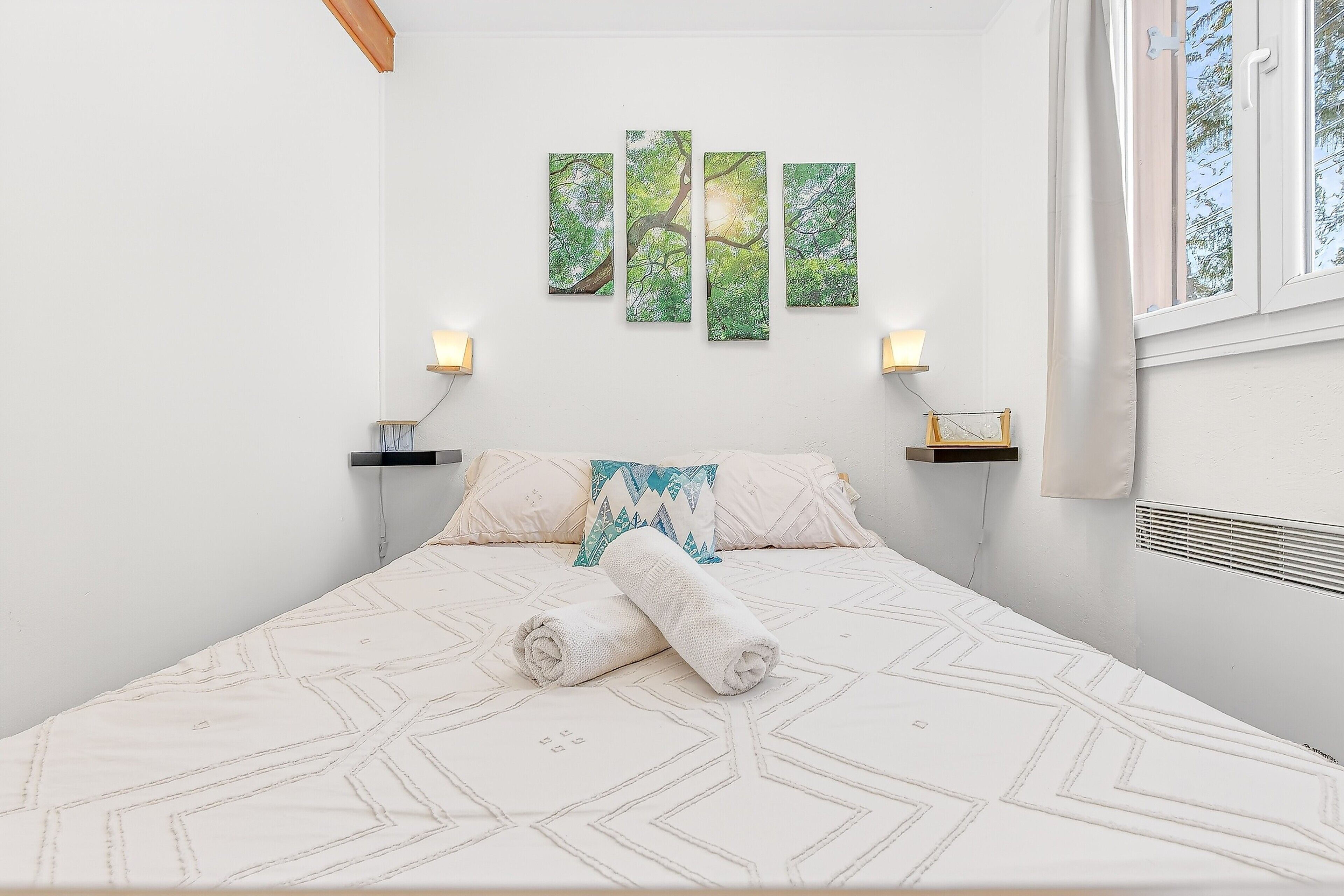 Un lit bien fait, recouvert d’une couvre-lit à motifs géométriques, une pile de serviettes enroulées et un mur orné de trois tableaux encadrés représentant des paysages naturels.
