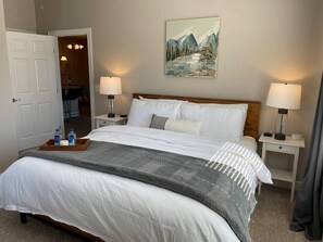 A neatly made bed with white linens, two bedside tables with lamps, a painting of a mountain landscape, and a wooden nightstand with a tray of toiletries. - 2BR Water Views W D Walkable Pool Gym (Westerville)