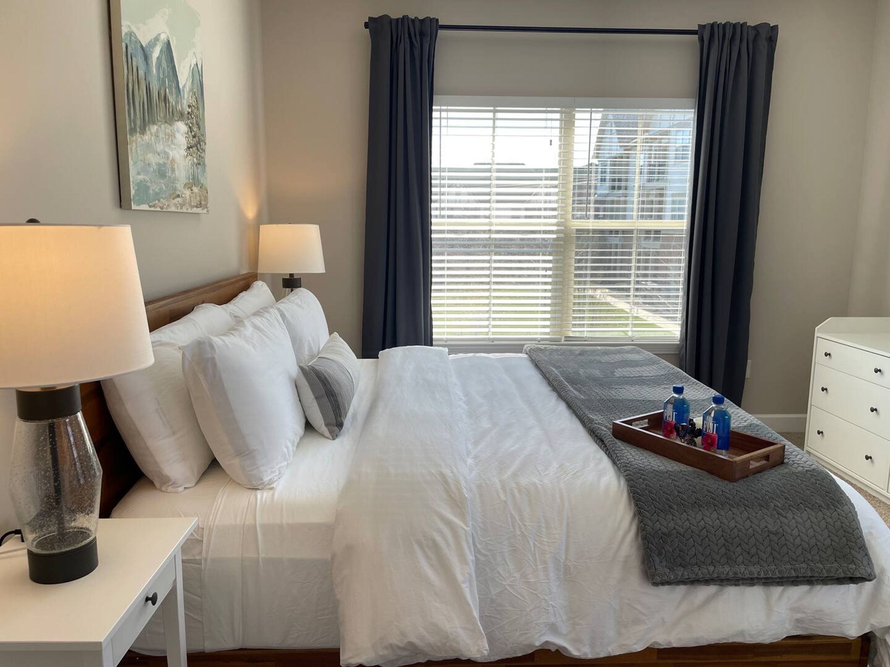 Ein Hotelzimmer mit Bett, zwei Nachttischlampen, einem Bild an der Wand, einem Fenster mit Vorhängen und einem Tablett mit Flaschen und Früchten auf dem Bett.
