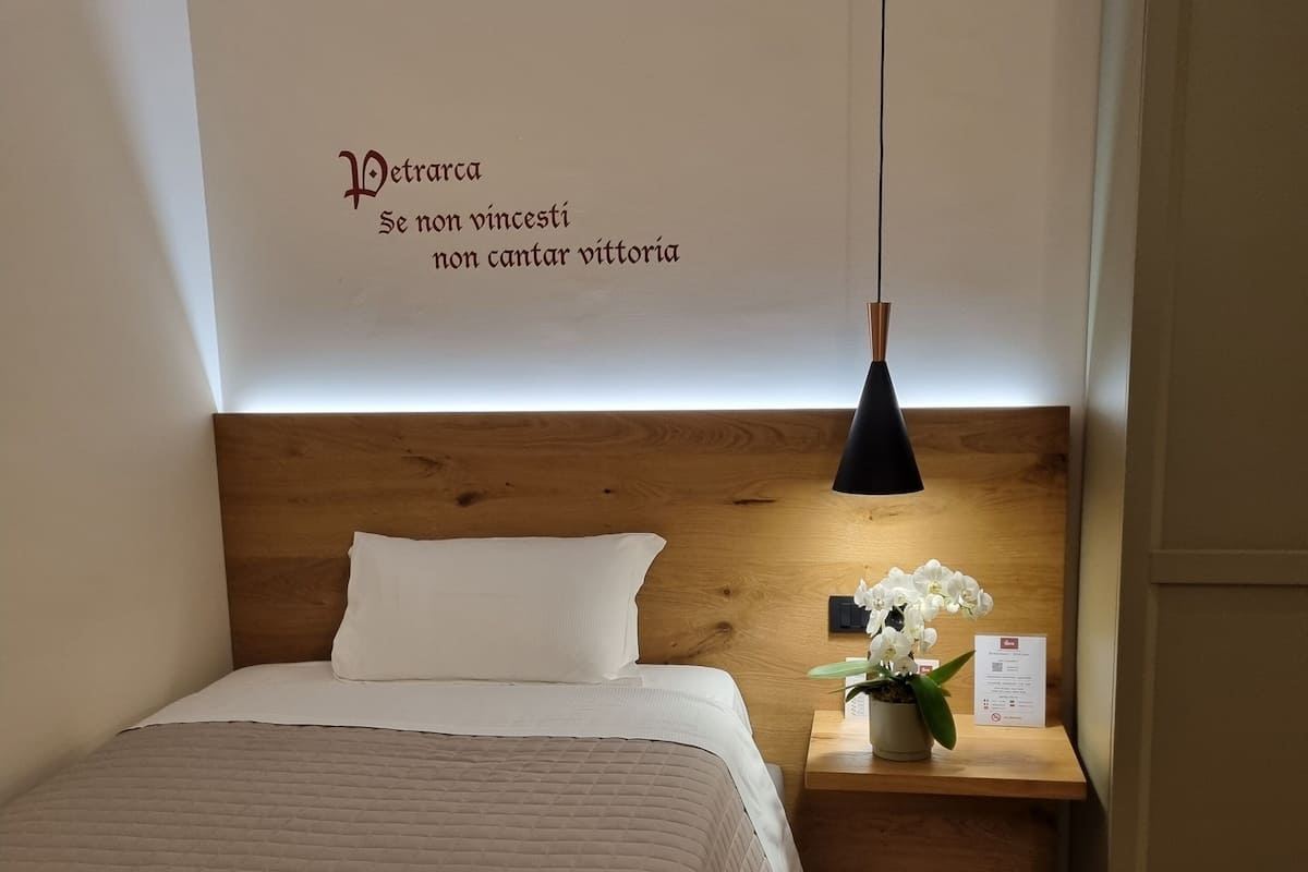 Een net opgemaakt bed met een wit kussen en een opgevouwen handdoek, een nachtkastje van hout met een potorkidee, en een muur met een citaat in Italiaans.