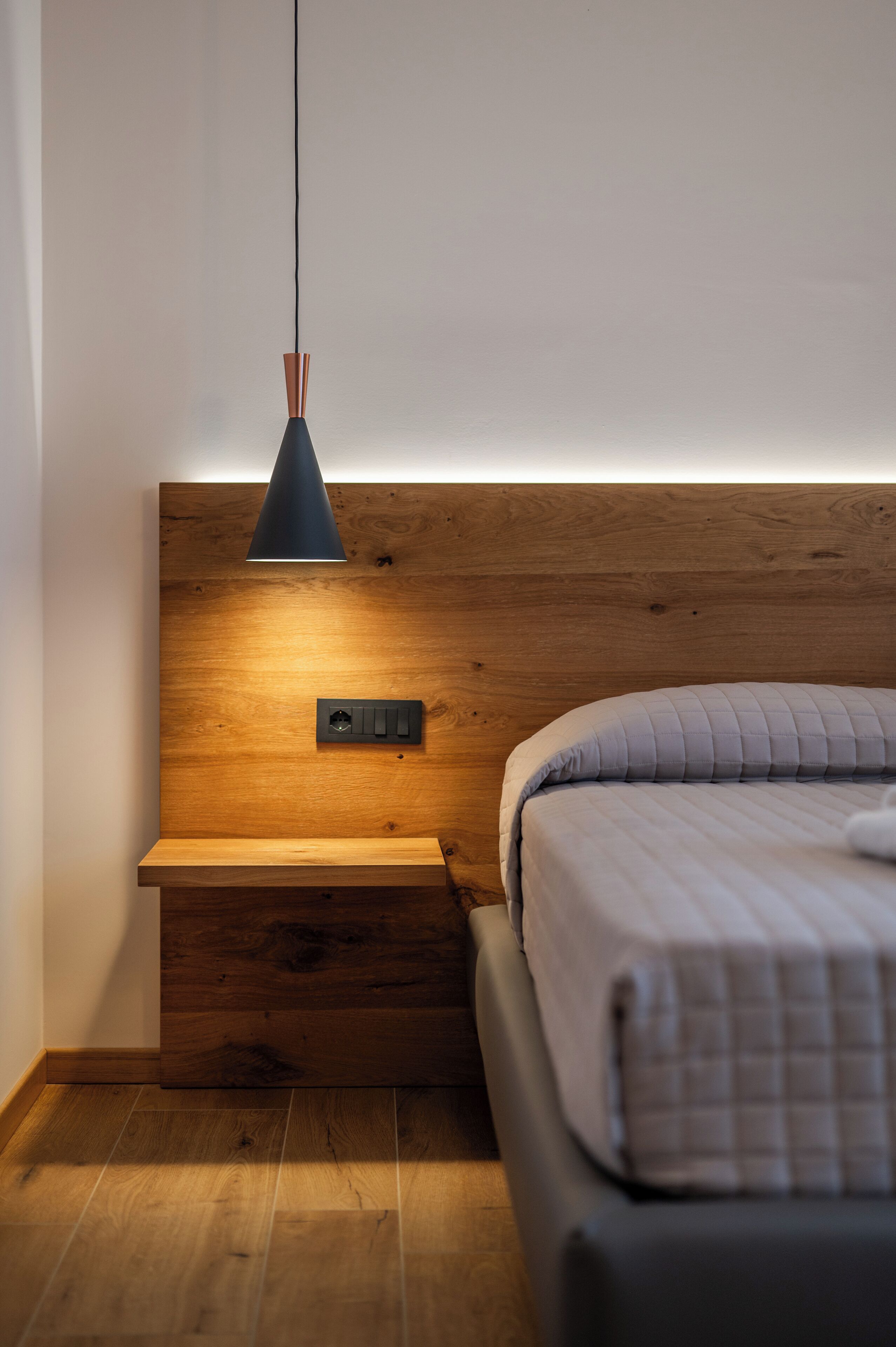 Une chambre à coucher avec une tête de lit en bois, une table de chevet, un lit recouvert d’une couverture matelassée et une lampe suspendue.