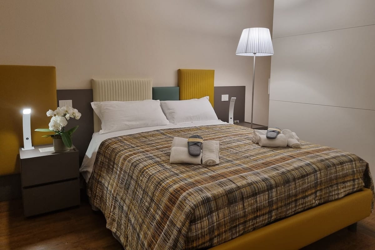 Een net opgemaakt bed met een ruitdessin sprei, een bedlampje en een nachtkastje met een vaas witte bloemen.