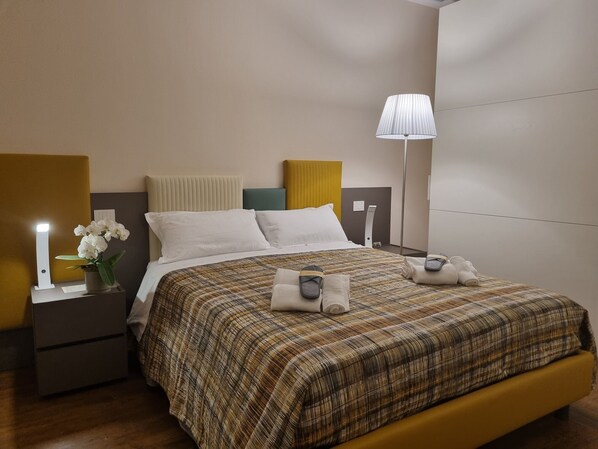 A neatly made bed with a plaid bedspread, a bedside lamp, and a nightstand with a vase of white flowers. - Aura - Dimore ad Arquà Petrarca (Arquà Petrarca)