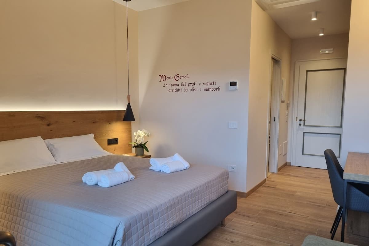 Een netjes opgemaakt bed met kussens en opgevouwen handdoeken, een nachtkastje met een lamp en bloemen, en een wand met een decoratief bord.