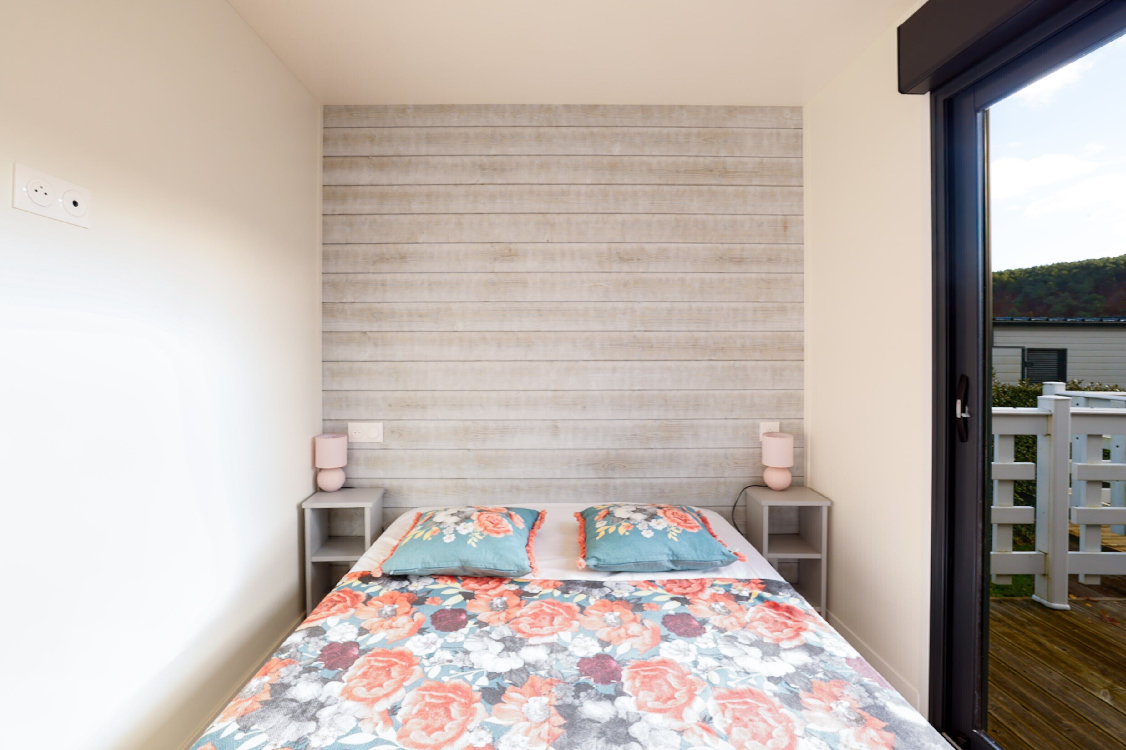 Ein Schlafzimmer mit Bett, Holz-Kopfteil, Nachttischen und Blick durch eine Glastür.