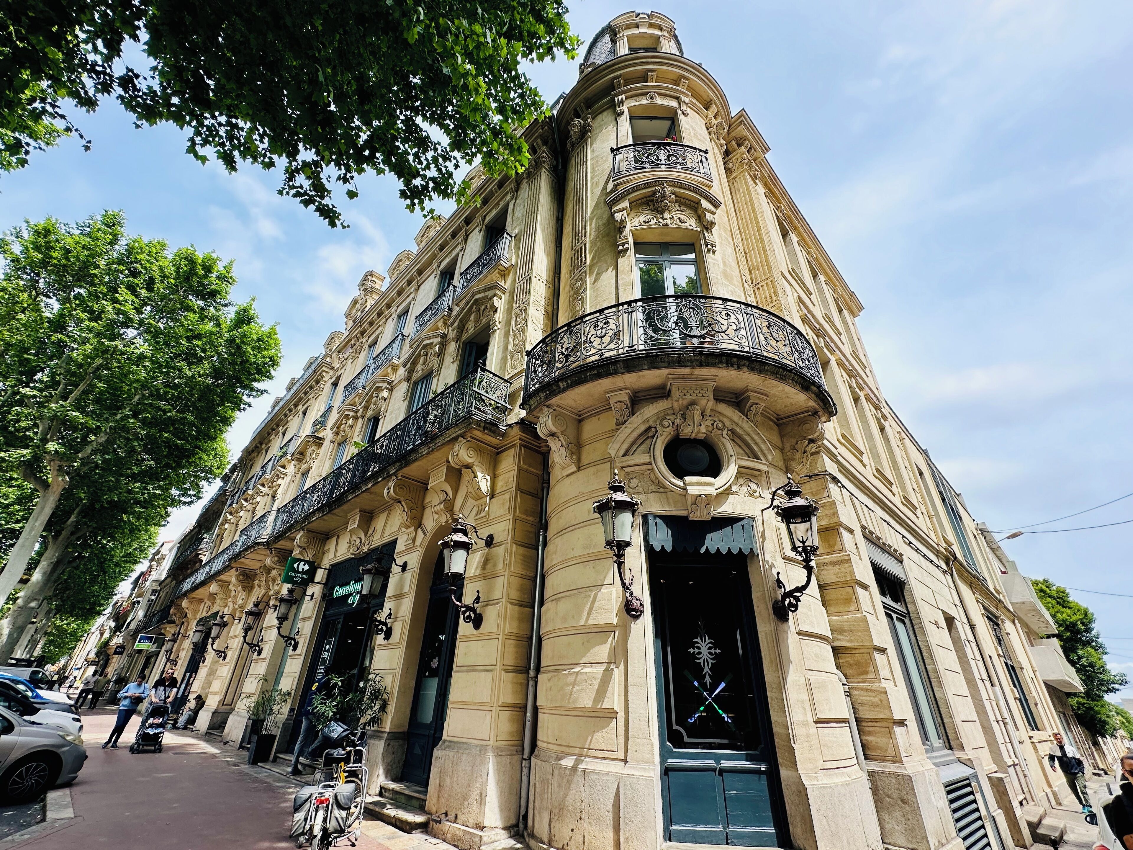 Un edificio histórico con arquitectura ornamentada, que incluye un balcón con intrincados detalles de hierro forjado, ventanas amplias y una entrada destacada con una puerta decorativa.