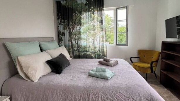 Een slaapkamer met een bed, kussens, een stoel, een raam en een televisie.
