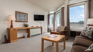 Un salon comprenant un bureau en bois, une télévision, un canapé et une grande fenêtre avec des rideaux.