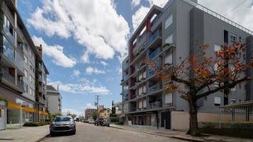 Un edificio residencial de varios pisos con balcones, un árbol con hojas otoñales y automóviles estacionados en la calle.