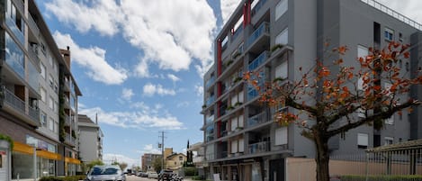 A multi-story residential building with balconies, a tree with autumn leaves, and parked cars on the street.