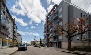 A multi-story residential building with balconies, a tree with autumn leaves, and parked cars on the street.