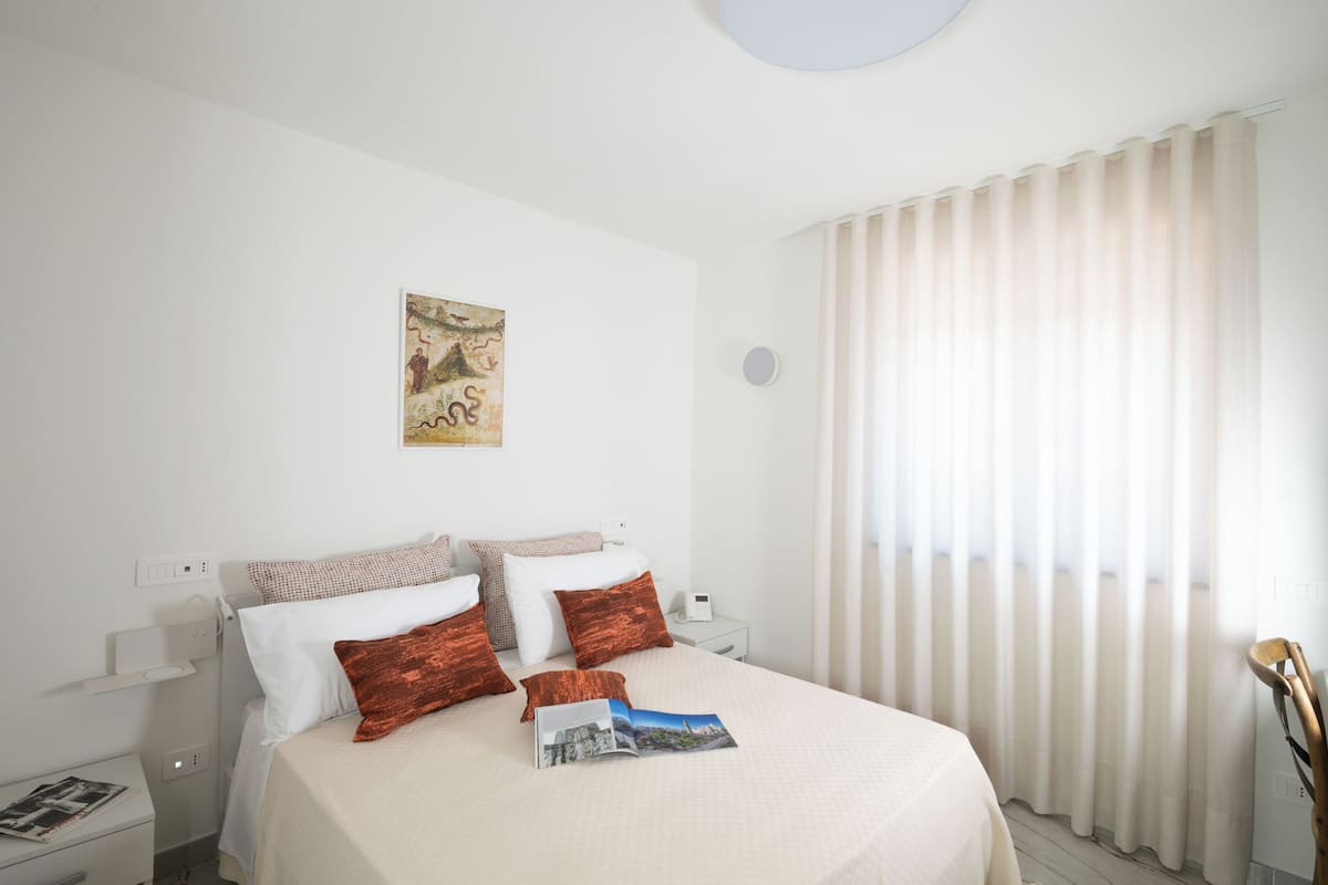 Une chambre d’hôtel bien rangée, avec un lit, des oreillers, une table de chevet, une horloge murale et une fenêtre avec des rideaux.