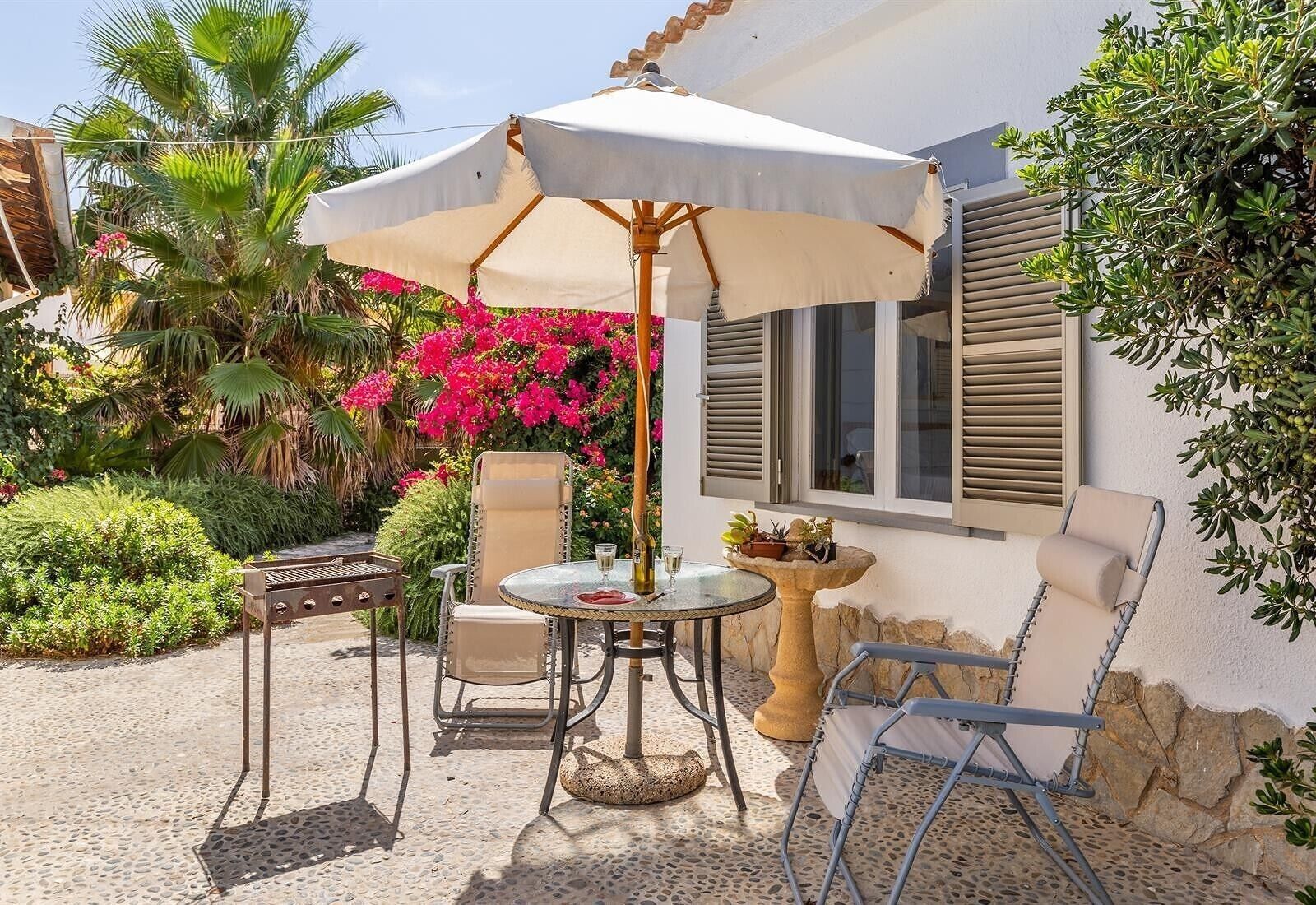Un pati exterior amb taula i cadires, una barbacoa, una banyera per a ocells decorativa i un para-sol blanc.