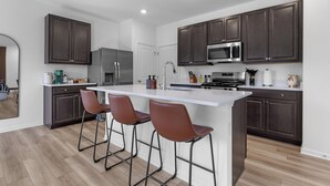 Une cuisine moderne avec des meubles en bois foncé, un îlot central blanc doté de tabourets, des appareils électroménagers en acier inoxydable et une grande fenêtre.