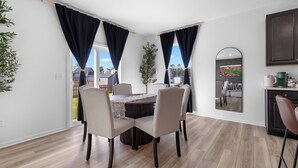 Un espace repas moderne avec une table ronde et quatre chaises, un coin cuisine doté de meubles sombres et une vue sur un quartier résidentiel grâce à de grandes fenêtres.