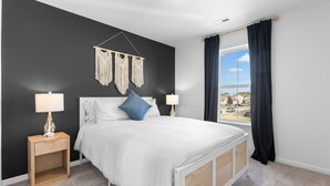 Une chambre à coucher avec un lit, deux tables de chevet avec des lampes, une décoration murale en macramé et une fenêtre avec des rideaux.