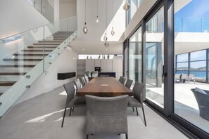 A modern interior with a large wooden table surrounded by chairs, a staircase, and large windows offering a view.