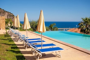 A poolside area with lounge chairs and umbrellas, a clear view of the sea, and a rocky hillside in the background.