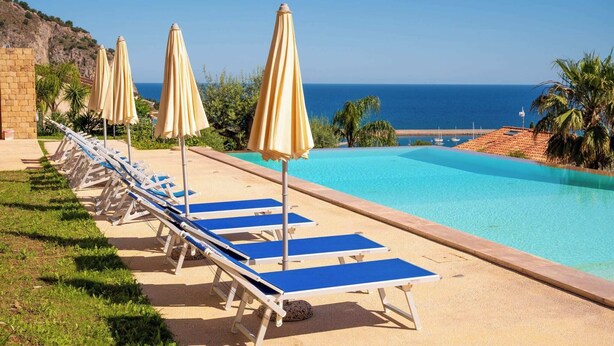 A poolside area with lounge chairs and umbrellas, a clear view of the sea, and a rocky hillside in the background. - CefalHome - MAGARA (Sicilia)