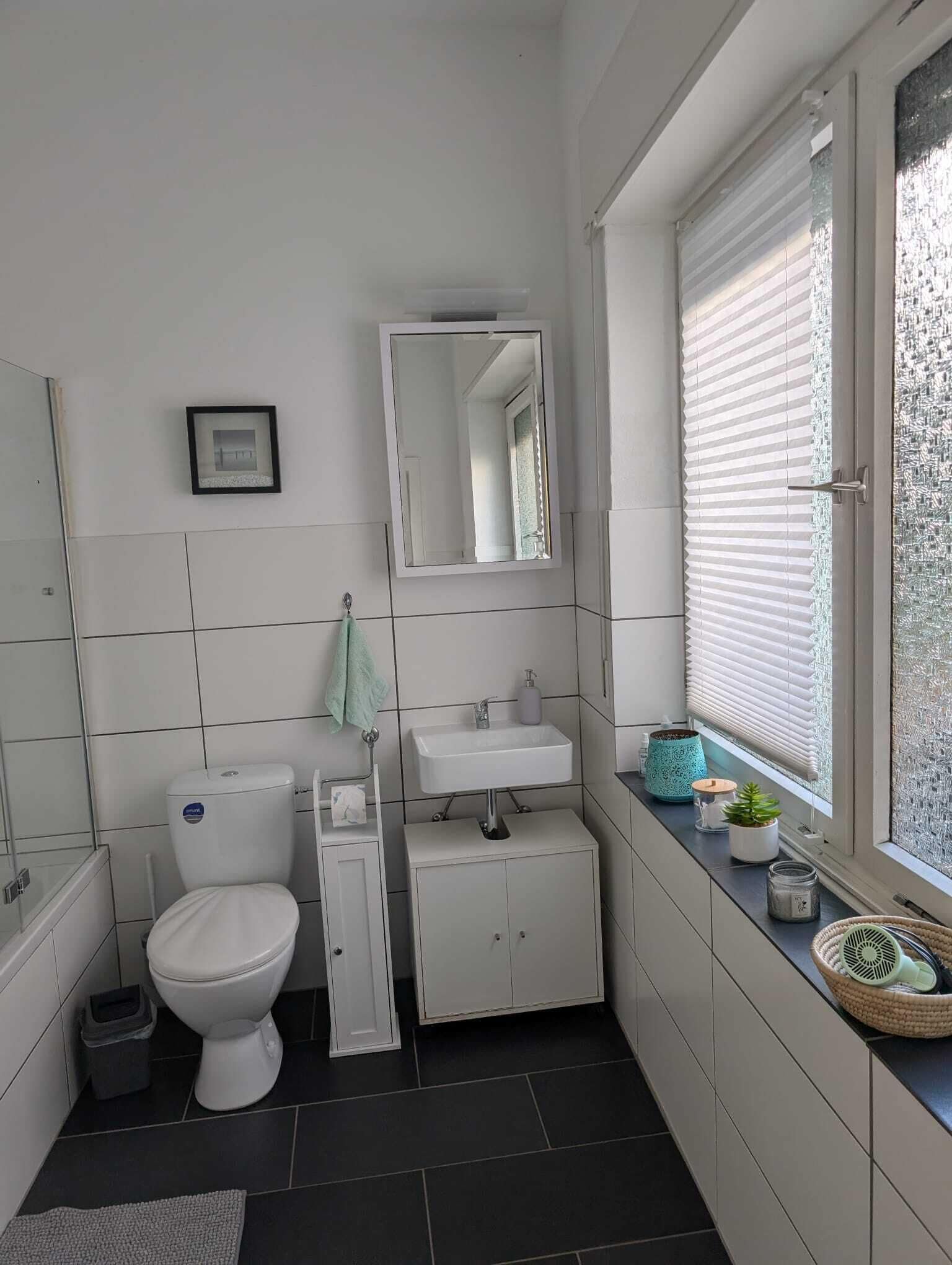 Ein modernes Badezimmer mit Toilette, Waschbecken und Spiegel. Ein Fenster mit Jalousien und ein kleiner Topfpflanze auf dem Fensterbrett.