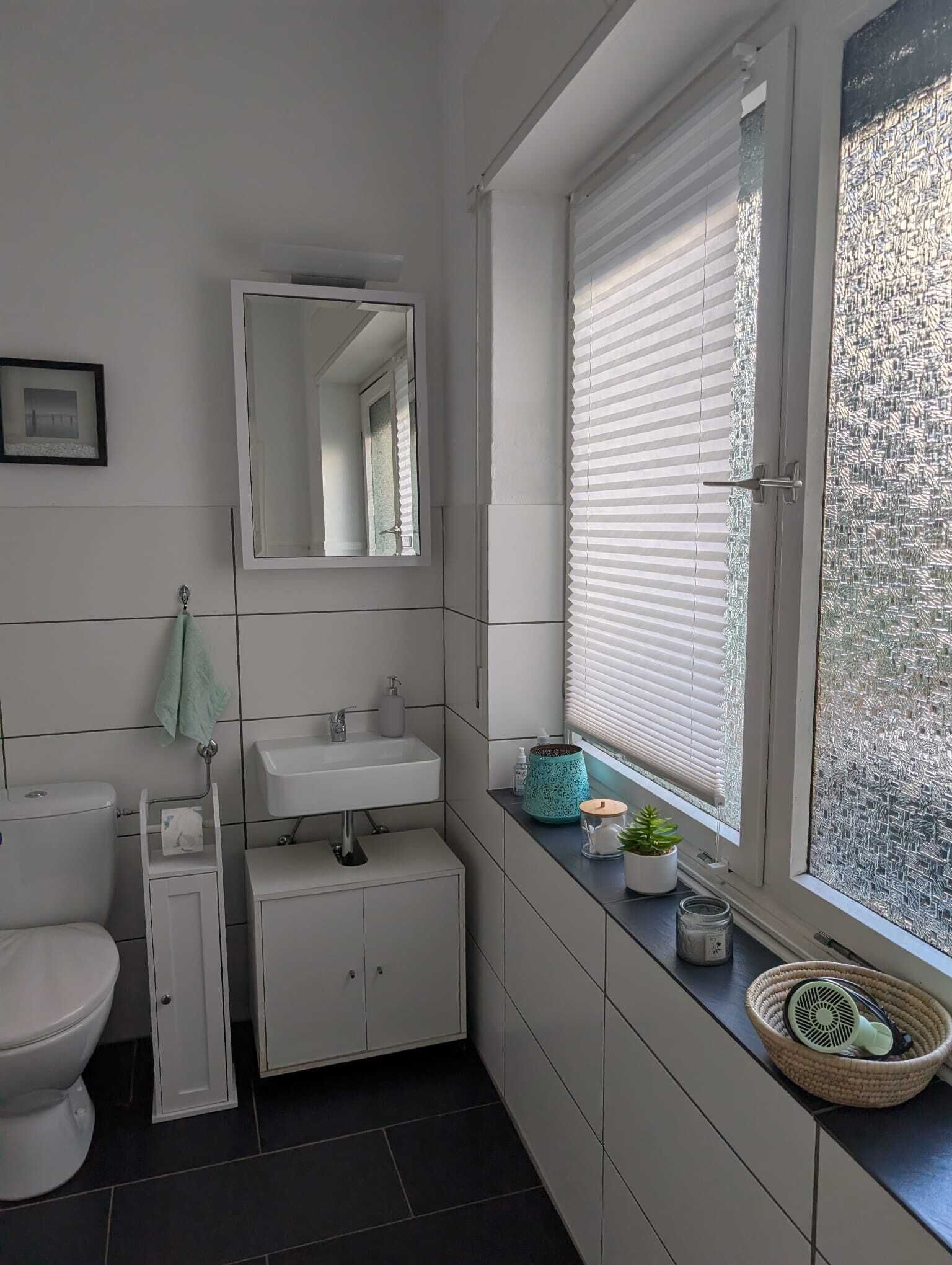 Uma casa de banho moderna com sanita, lavatório e espelho. Tem uma janela com estores e vista para o exterior.