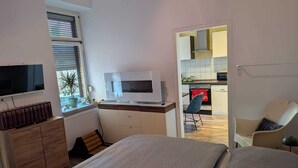 Een knus slaapkamer met een bed, een televisie, een bureau met een computer, en uitzicht op een keuken en eethoek.