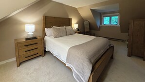A bedroom with a sloped ceiling, a bed with a wooden headboard, bedside tables with lamps, a window, and a wardrobe.