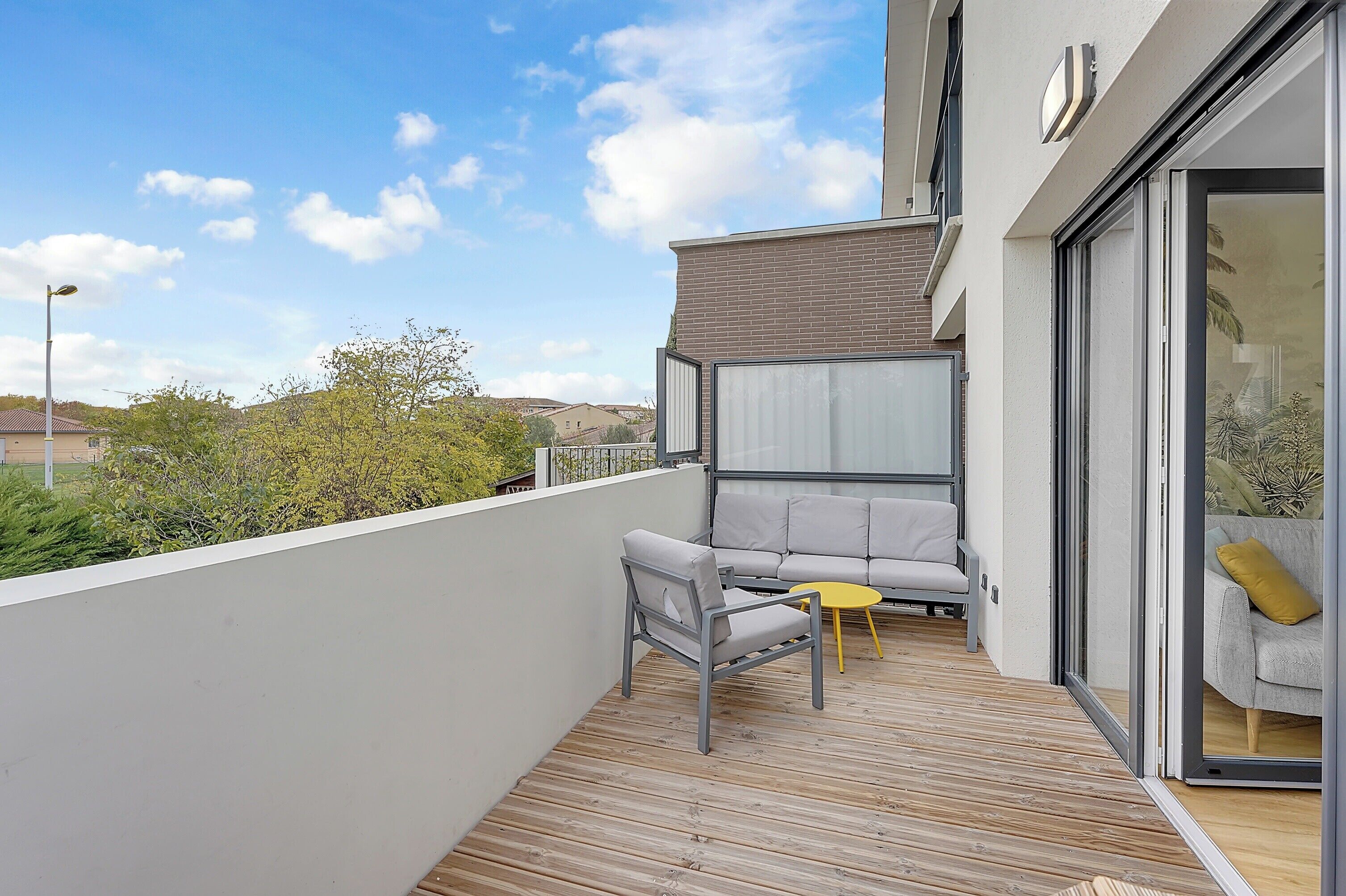 A balcony with a grey sofa, a small yellow table, and a sliding glass door leading to a room with a grey sofa and a yellow pillow.