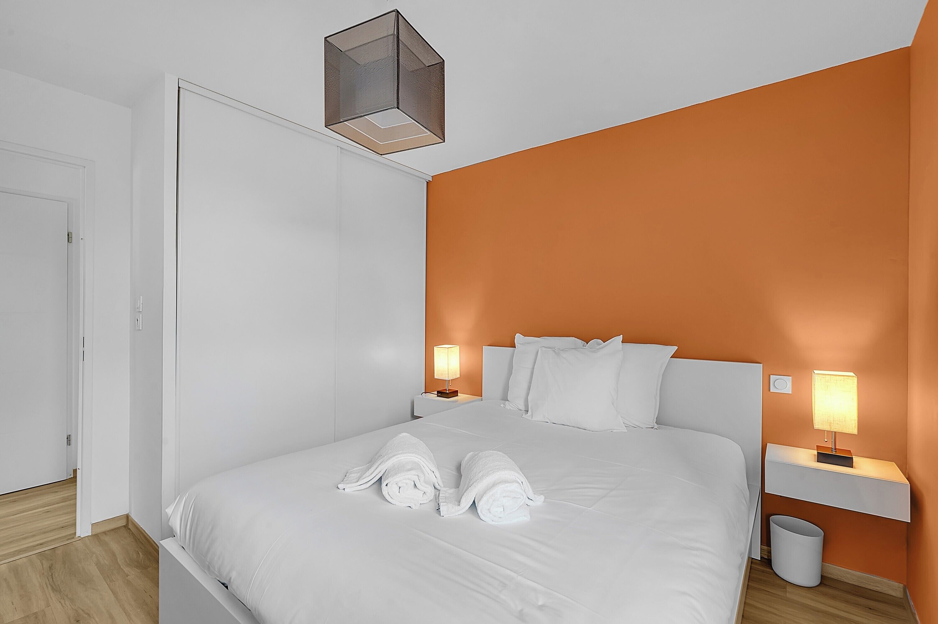 Een keurig opgemaakt bed met witte lakens en twee opgerolde handdoeken, een nachtkastje met een lamp, en een kleine prullenbak.