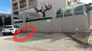 Ein durch einen Zaun gesicherter Parkbereich mit einem weißen Auto vor einer Skulptur eines Baumes und einem Gebäude mit Balkonen sowie einem Schild mit der Aufschrift 'LE MAIL BIA'.
