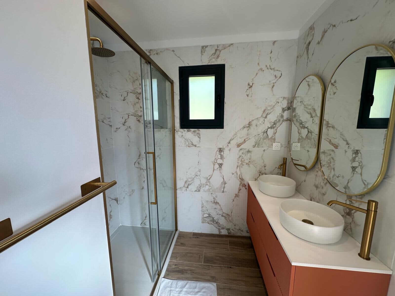 Une salle de bain moderne avec des murs en marbre, une douche en verre et deux vasques avec des robinets dorés.