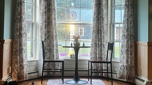 Ein Erker mit gemusterten Vorhängen, ein Tisch mit einer Vase und zwei Stühle.