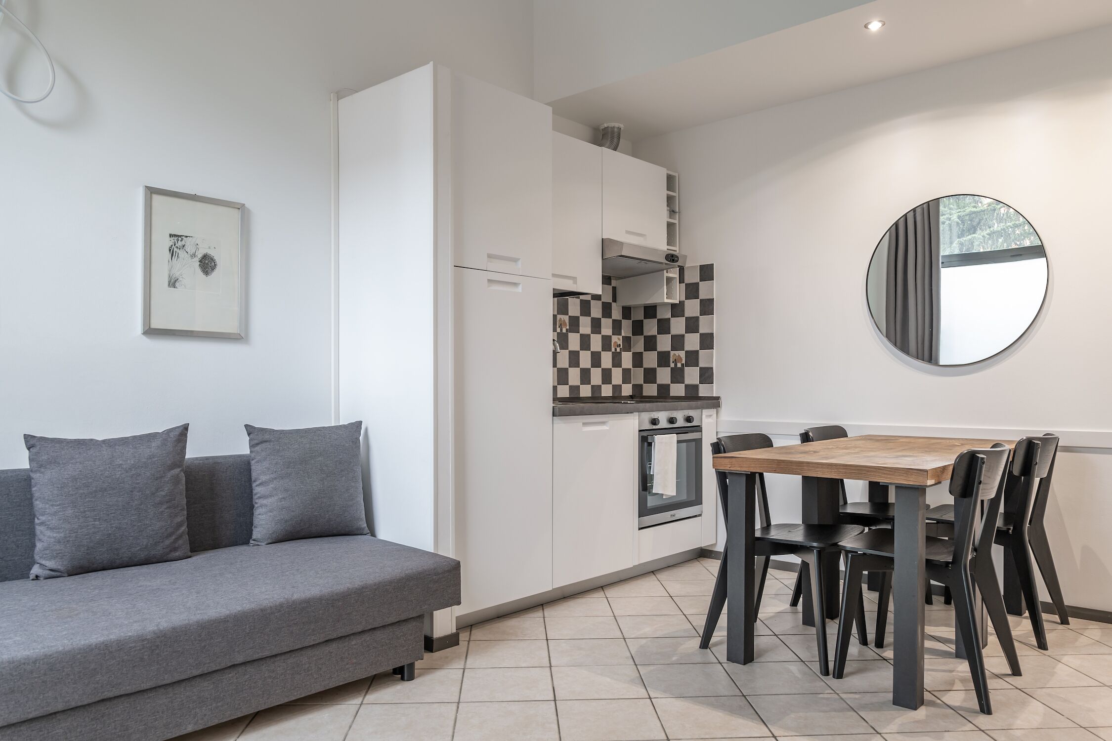 A modern kitchen with white cabinets, a checkered backsplash, a round mirror, and a dining area with a wooden table and black chairs.