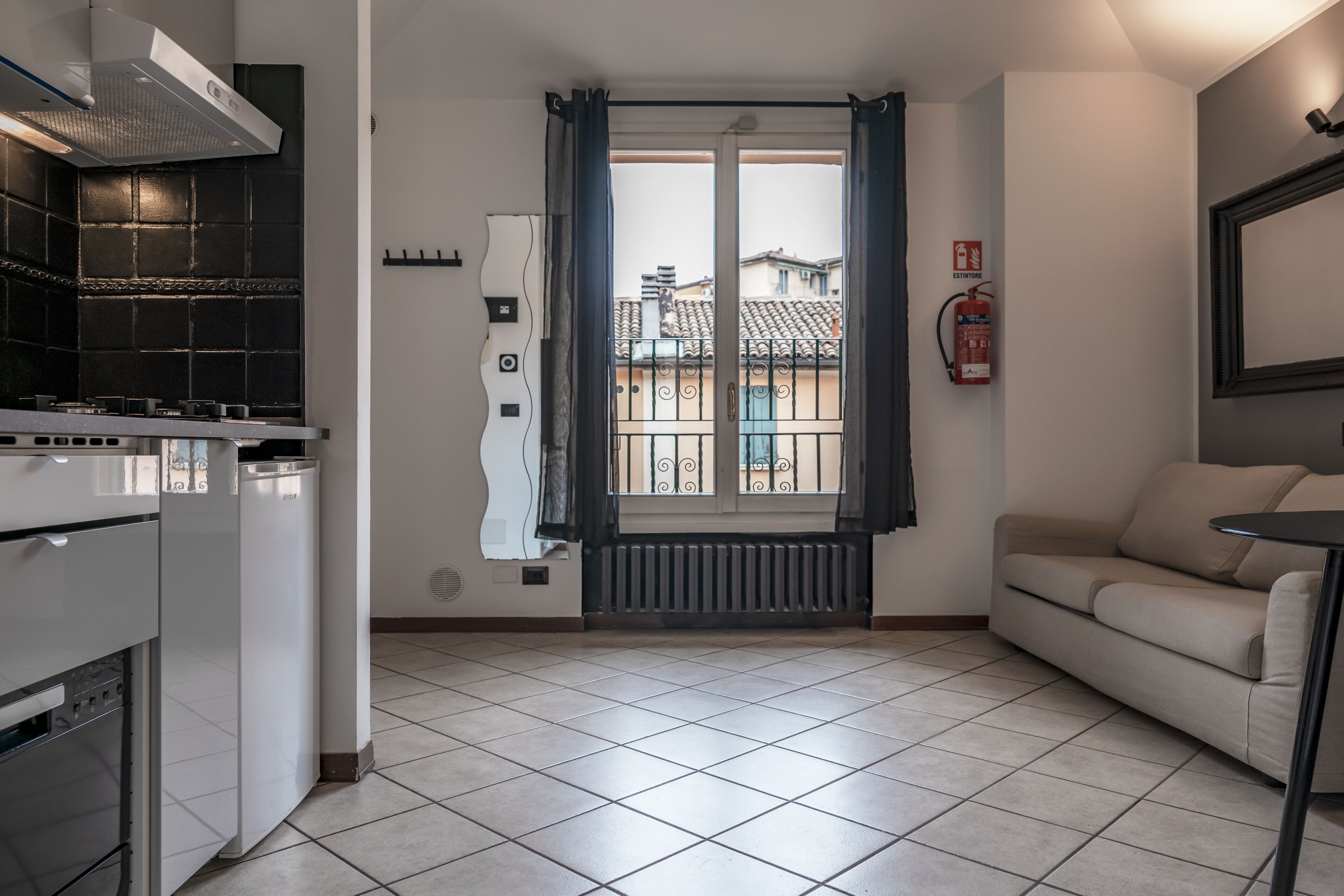 Eine kompakte Küche mit Herd, Backofen und moderner Dunstabzugshaube. Ein Fenster bietet Blick auf umliegende Gebäude, und ein Feuerlöscher ist an der Wand angebracht.