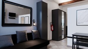 Een moderne hotelkamer met een donkergrijze bank, een grote spiegel, een flatscreen televisie, een brandblusser en een zithoek met een tafel en stoelen.