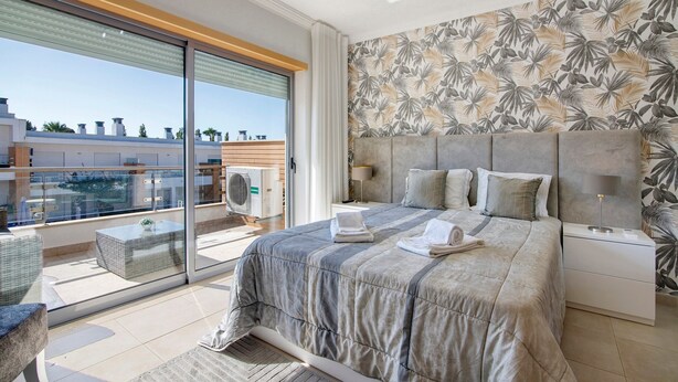 Une chambre avec un grand lit, une table de chevet, une lampe, une fenêtre donnant sur des immeubles et un balcon avec une table et des chaises.
